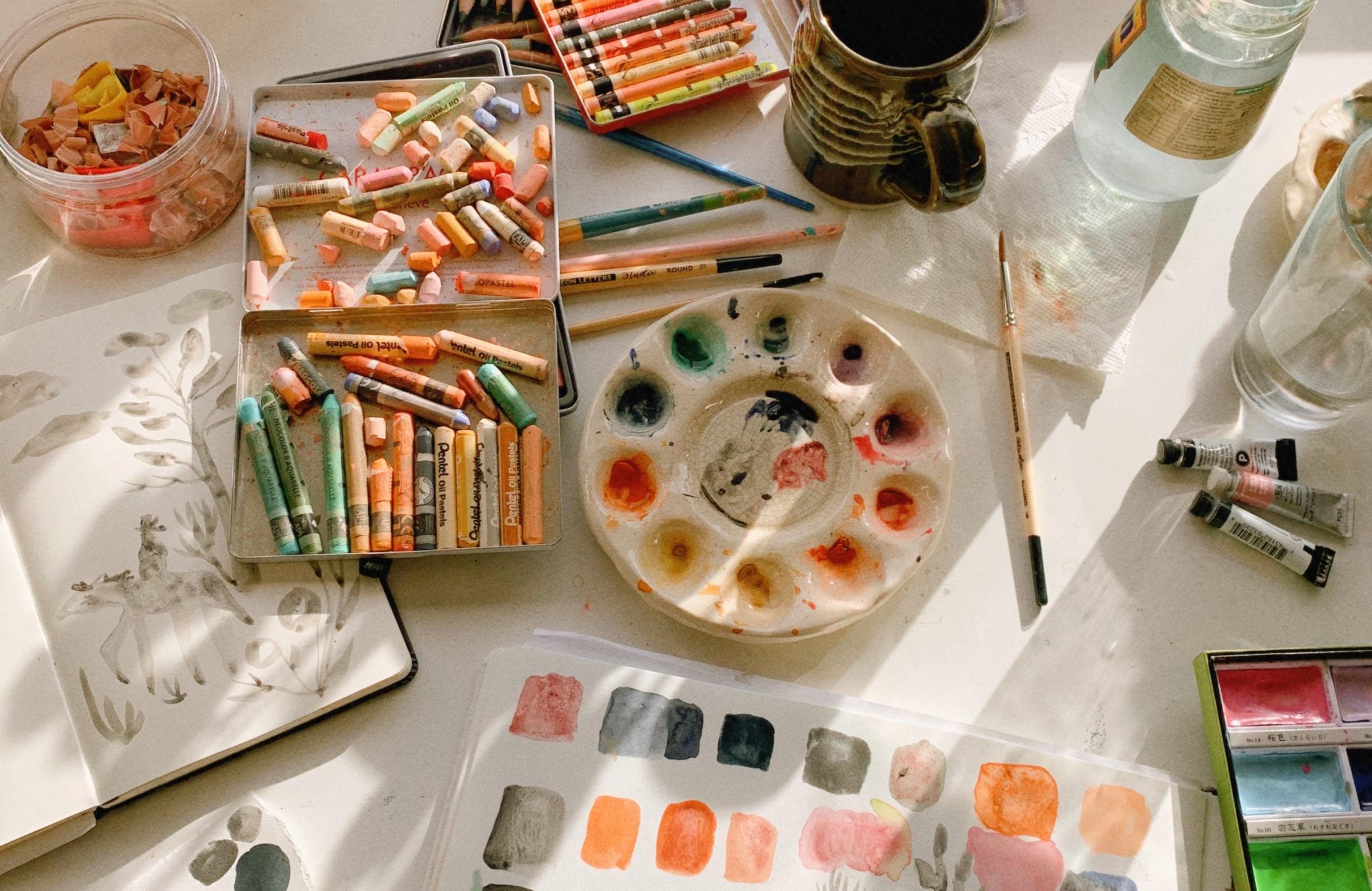 An array of paint and painting supplies on a work table with open sketch books