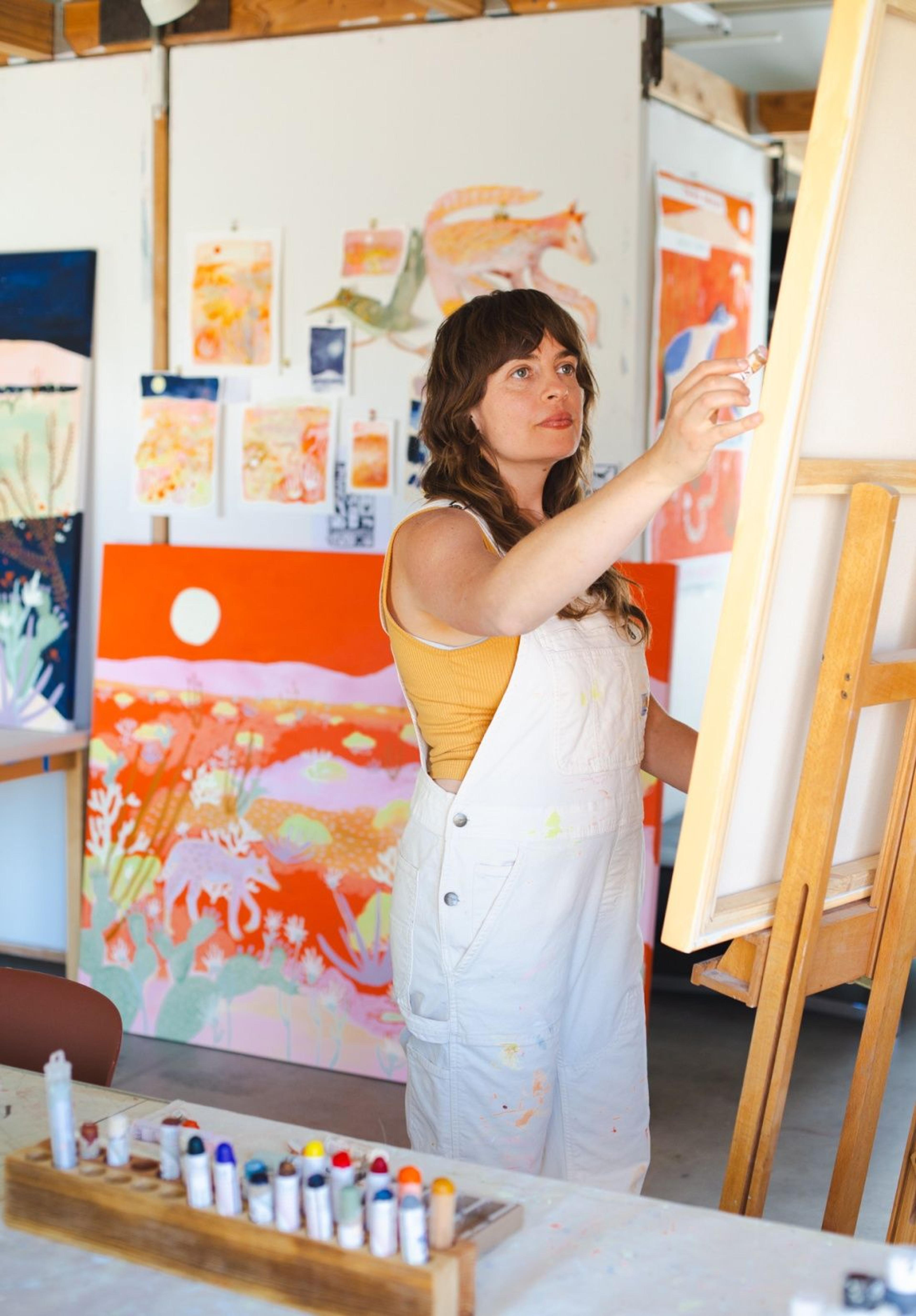 Artist Melissa Lakey painting at her easel at her studio in Joshua Tree with colorful paintings surrounding her