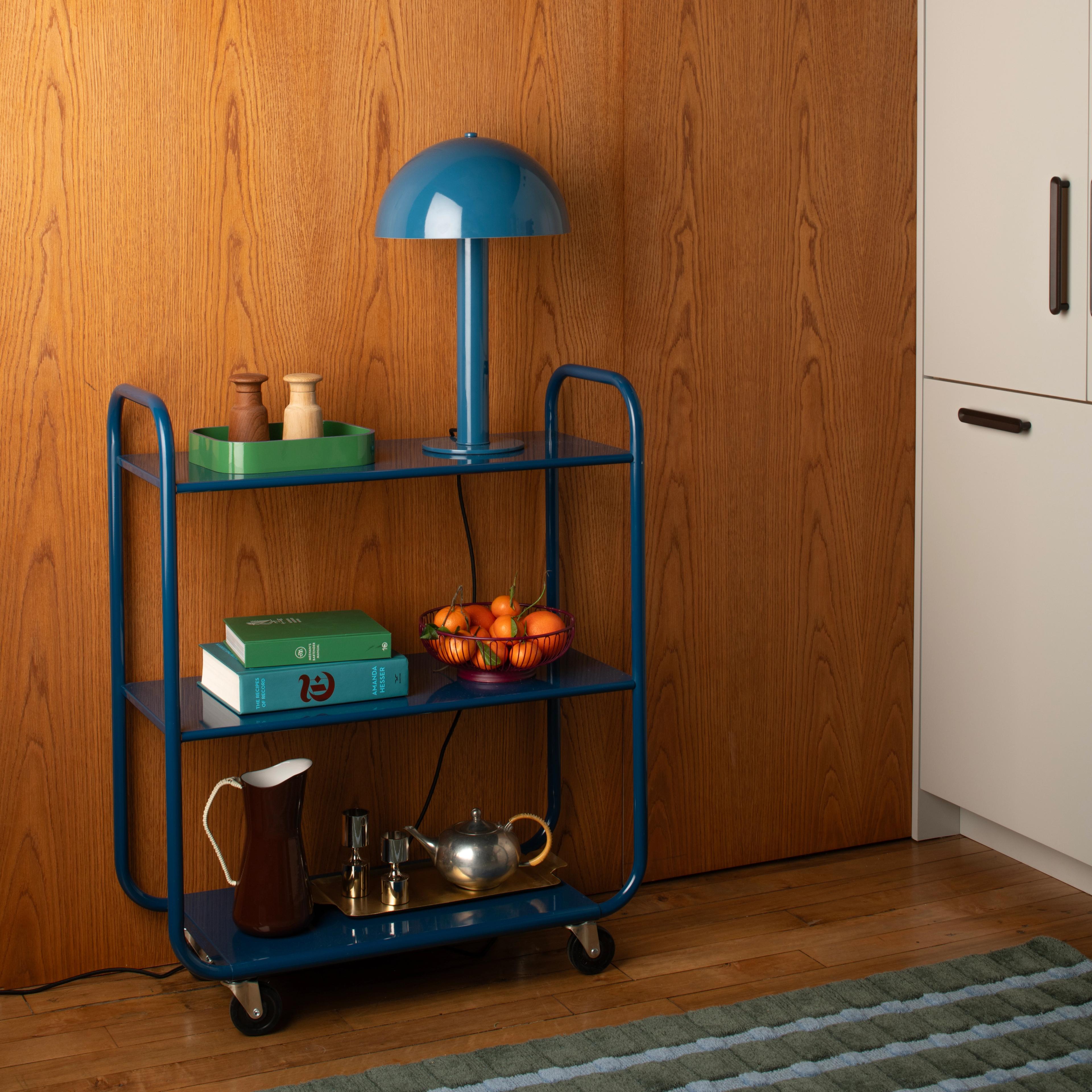 Utility cart with blue mushroom shaped lamp, stacked cook books and serving pitchers on bottom shelf