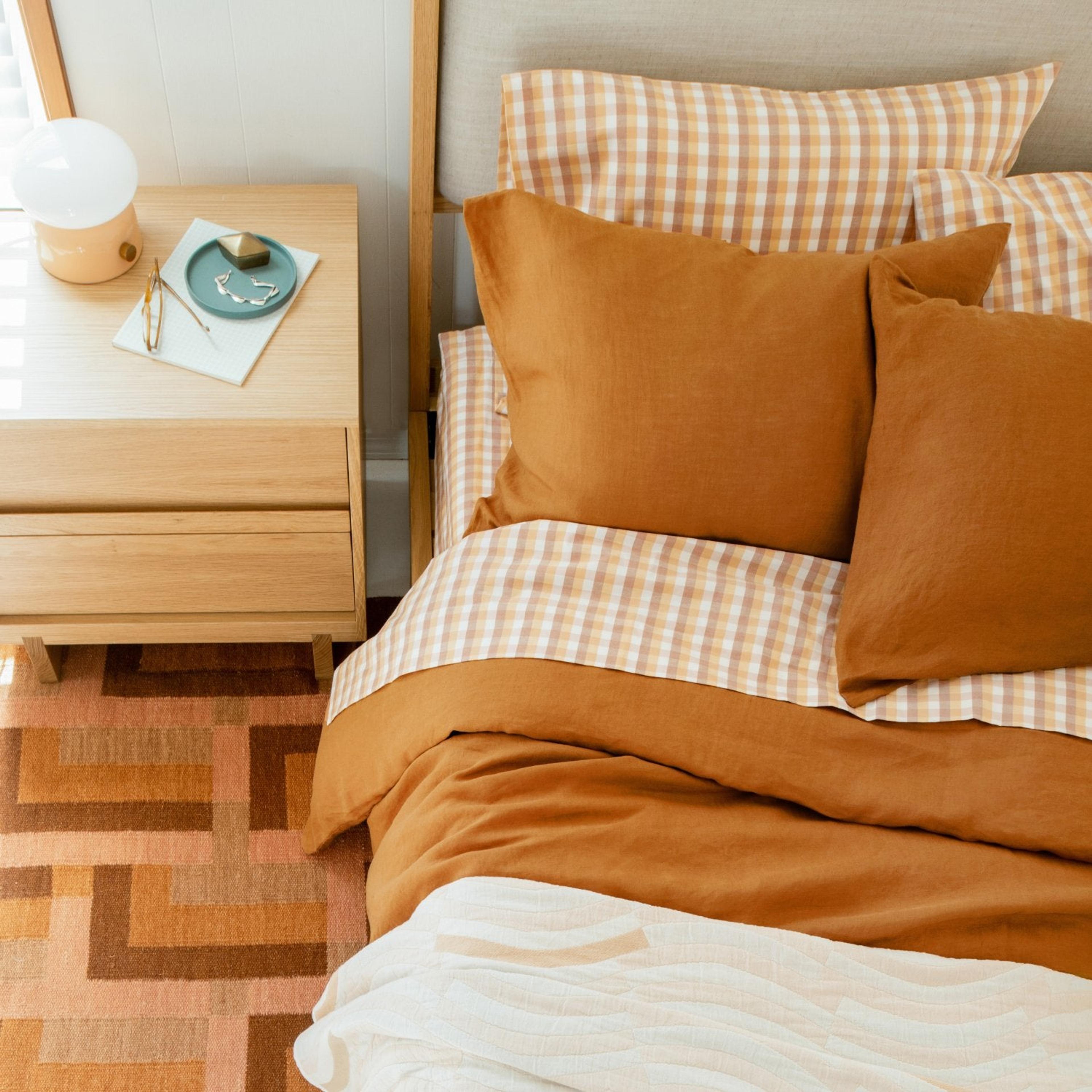 Orange hued bedset from above the bed with orange and checkered white/orange sheets, wooden side table with small lamp on top