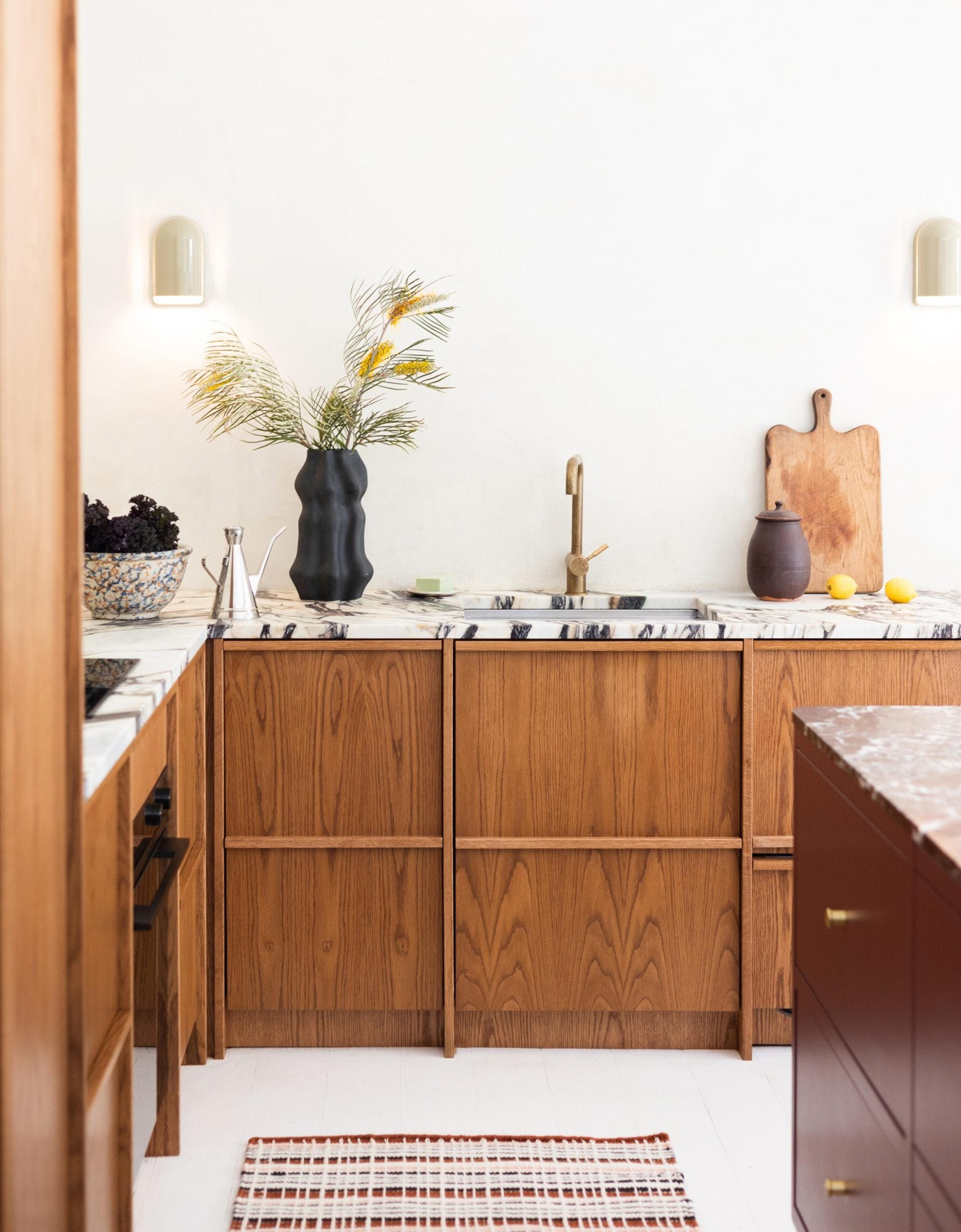 Kitchen with wood cabinets, marble countertops, vase with flowers next to sink, white wall with small white pendant lights